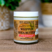 Jar of Mion Artisan Soap Whipped Shea Butter Pink Grapefruit on a wooden surface with a blurred green plant in the background.