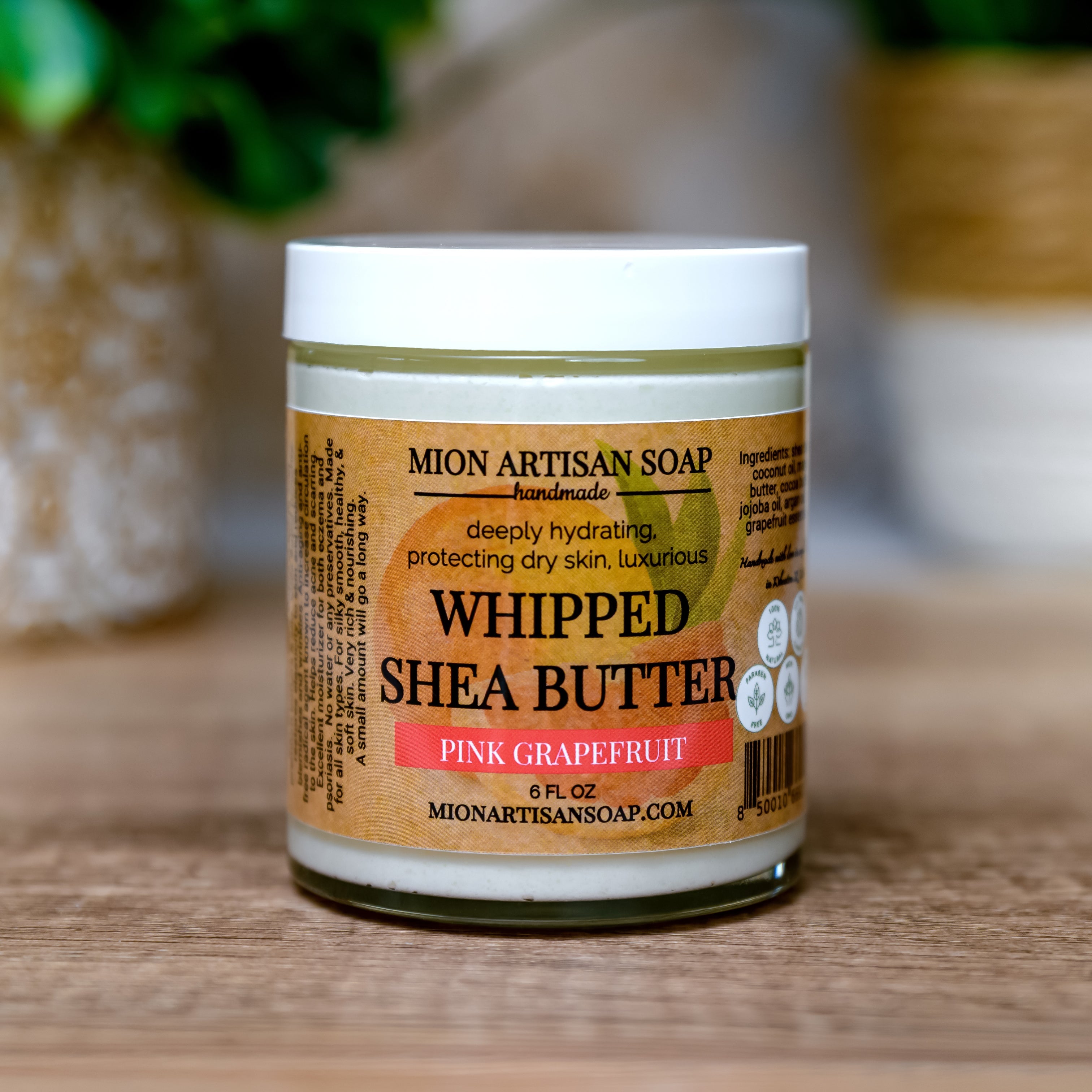Jar of Mion Artisan Soap Whipped Shea Butter Pink Grapefruit on a wooden surface with a blurred green plant in the background.