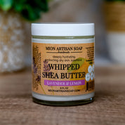 Jar of Mion Artisan Soap whipped shea butter with a blurred green plant in the background