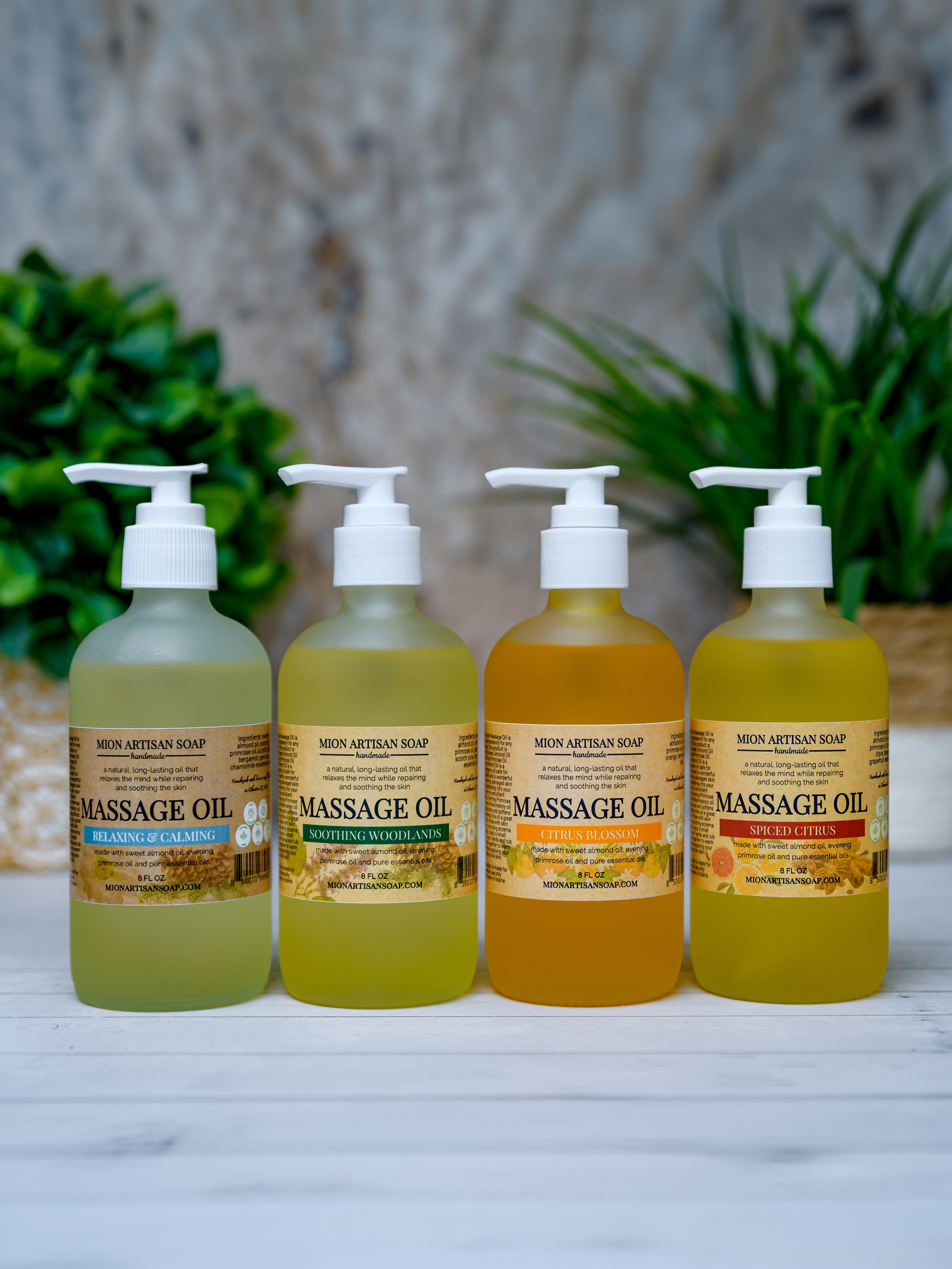Four bottles of massage oil with different colored oils on a white surface with a blurred natural background.