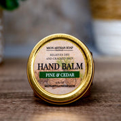Hand balm container labeled 'Pine & Cedar' on a wooden surface with a blurred natural background.