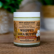 Jar of Mion Artisan Soap Whipped Shea Butter on a wooden surface with a blurred green plant background.