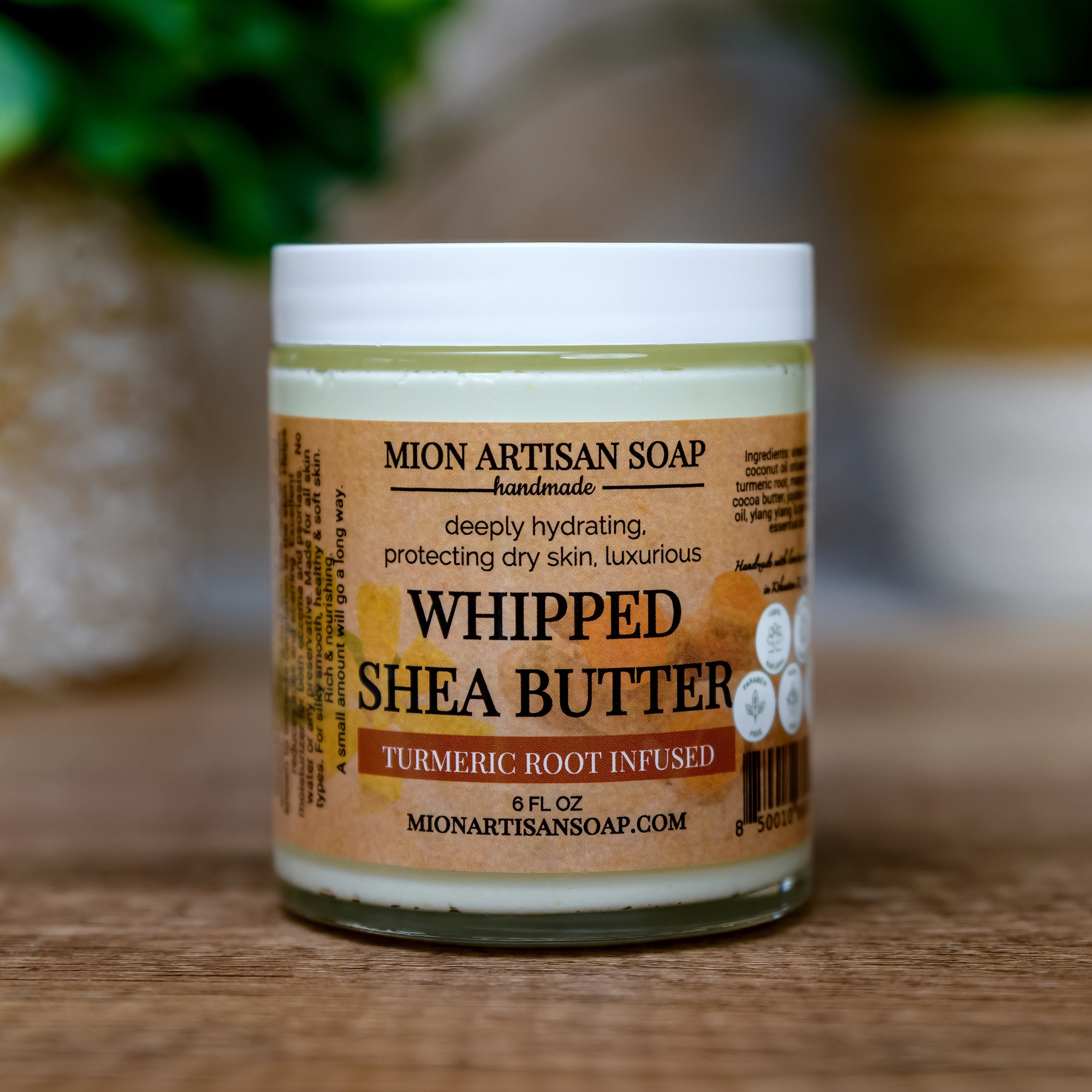 Jar of Mion Artisan Soap Whipped Shea Butter on a wooden surface with a blurred green plant background.