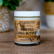 Jar of Mion Artisan Soap Whipped Shea Butter on a wooden surface with a blurred green plant in the background