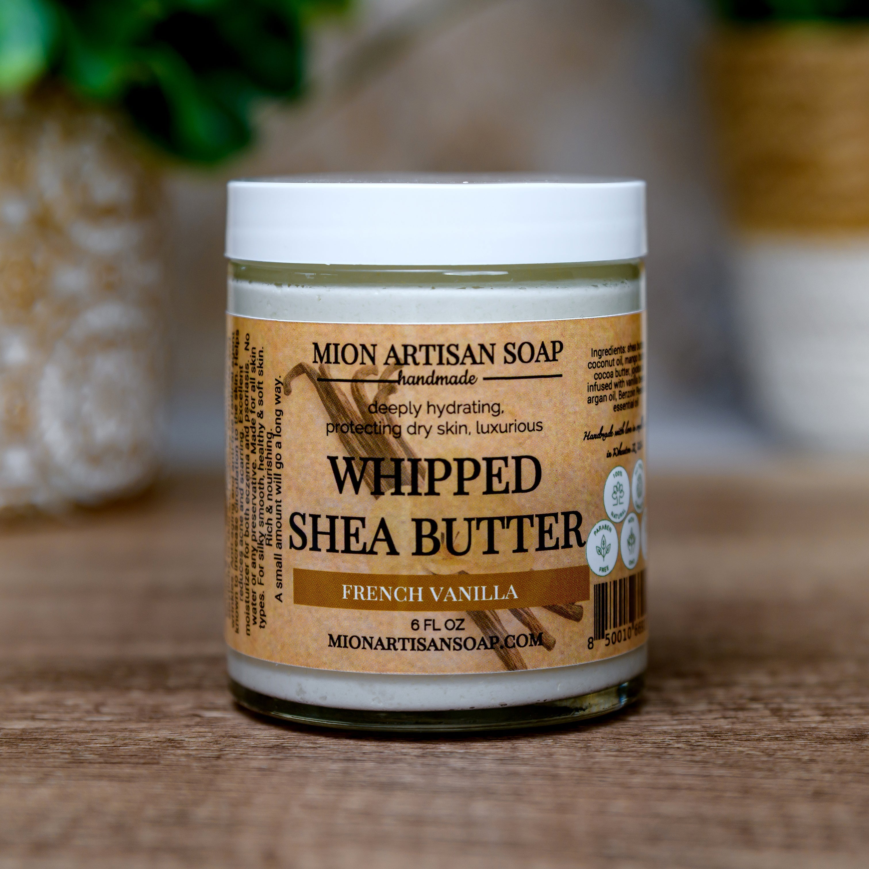 Jar of Mion Artisan Soap Whipped Shea Butter on a wooden surface with a blurred green plant in the background